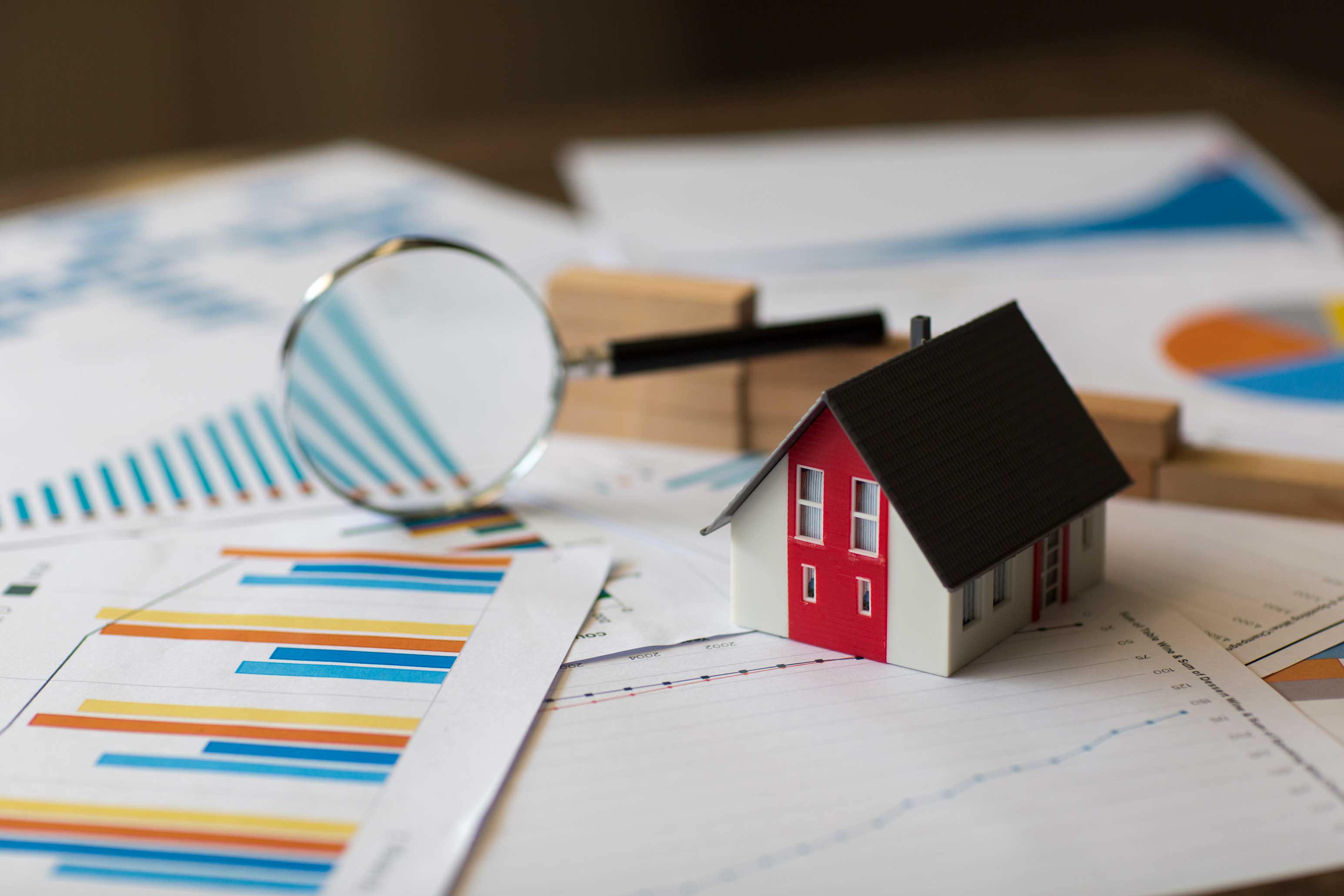 Photo of a miniature house on a set of papers with financial graphs and a magnifying glass in the background 